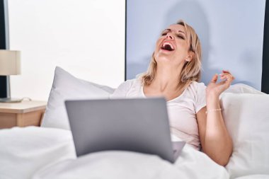Young blonde woman having video call sitting on bed at bedroom