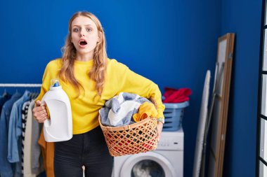 Young caucasian woman holding laundry basket and detergent bottle afraid and shocked with surprise and amazed expression, fear and excited face. 