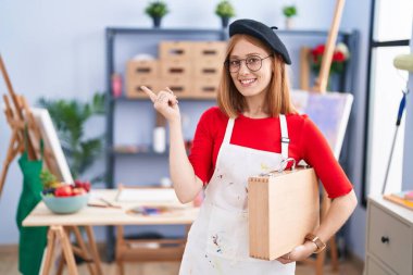 Young redhead woman at art studio holding art case with a big smile on face, pointing with hand finger to the side looking at the camera. 