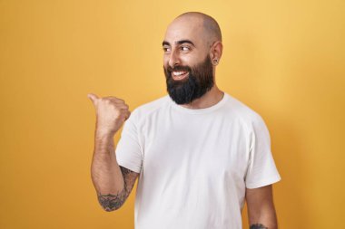 Young hispanic man with beard and tattoos standing over yellow background smiling with happy face looking and pointing to the side with thumb up. 