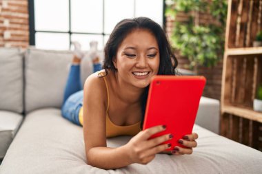 Young chinese woman using touchpad lying on sofa at home