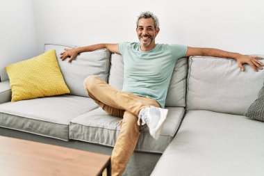 Orta yaşlı, kır saçlı, gülümseyen bir adam. Evdeki kanepede oturuyor.