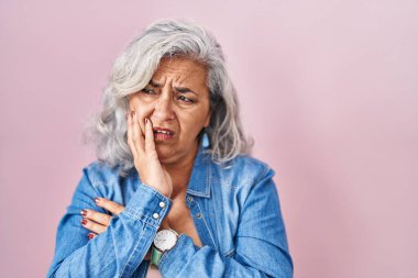Pembe arka planda duran gri saçlı orta yaşlı bir kadın. Elleri ağızda, tırnaklarını yerken gergin ve stresli görünüyor. anksiyete. 