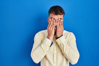 Handsome hispanic man standing over blue background rubbing eyes for fatigue and headache, sleepy and tired expression. vision problem 