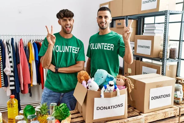 Young gay couple wearing volunteer t shirt at donations stand smiling with happy face winking at the camera doing victory sign with fingers. number two.