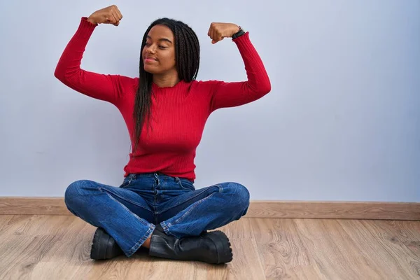年轻的非洲裔美国人 辫子坐在家里的地板上 露出胳膊肌肉 带着自豪的笑容 健身概念 — 图库照片