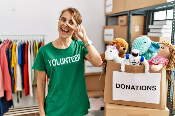 Beautiful caucasian woman wearing volunteer t shirt at donations stand doing peace symbol with fingers over face, smiling cheerful showing victory
