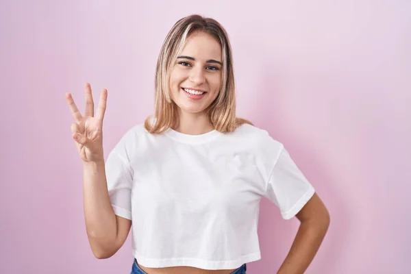 Молодая Блондинка Стоит Розовом Фоне Показывая Показывая Пальцами Номер Три — стоковое фото