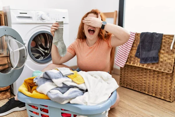 若い赤毛の女性は洗濯機に汚れた洗濯機を入れて笑顔と驚きのために目をカバー顔に手で笑っています 視覚障害の概念 — ストック写真