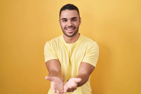 黄色の背景の上に立っている若いヒスパニック系の男が一緒に手の手のひらで笑顔を受信またはジェスチャーを与える 保持し保護する — ストック写真