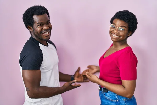 年轻的非洲裔美国夫妇站在粉红的背景上 带着开放的手 自然地微笑着走进来 — 图库照片
