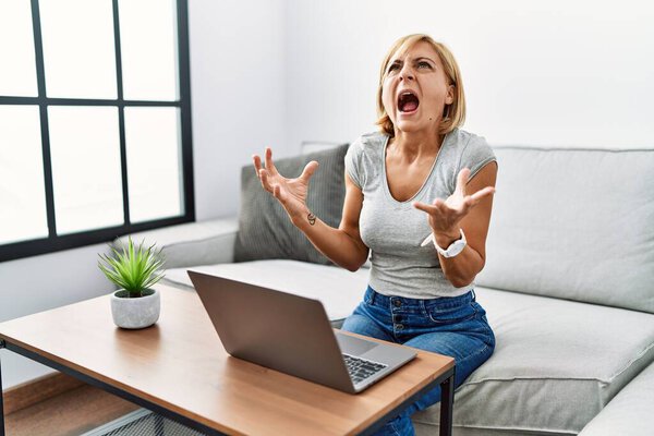 Блондинка средних лет использует ноутбук дома сумасшедшая и сумасшедшая кричит и кричит с агрессивным выражением лица и поднятыми руками. Концепция разочарования. 