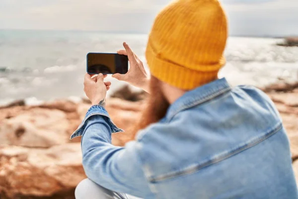 バックビューに立って若い赤毛男は海辺でスマートフォンで写真を撮る — ストック写真