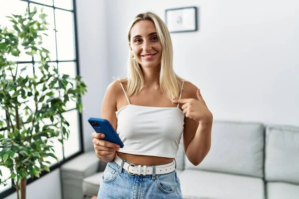 Young Blonde Woman Using Smartphone Typing Message Home Pointing Finger — Zdjęcie stockowe