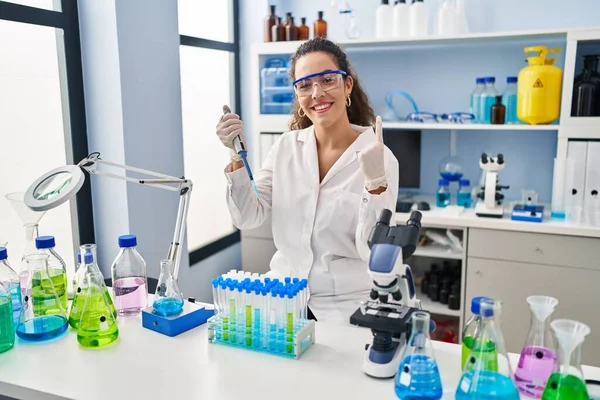 Young hispanic woman working at scientist laboratory smiling with an idea or question pointing finger with happy face, number one