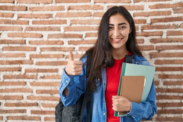 Young teenager girl wearing student backpack and holding books smiling happy and positive, thumb up doing excellent and approval sign