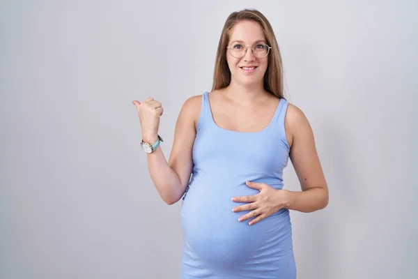 手と親指を上げて後ろを指差す白い背景の上に立つ若い妊婦は 自信を持って笑って — ストック写真