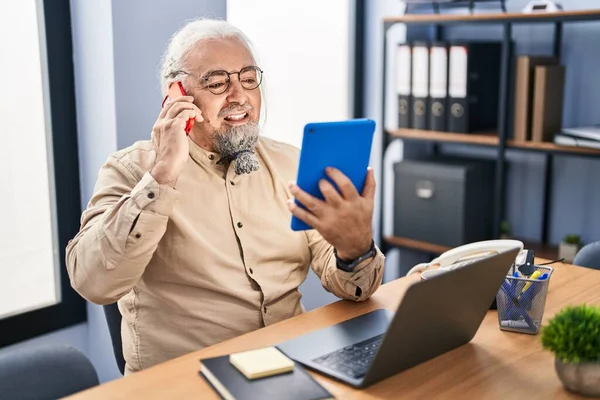 Middle age grey-haired man business worker talking on smartphone using touchpad at office