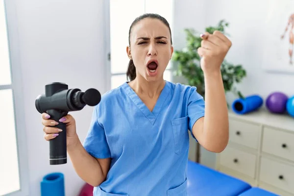 stock image Young physiotherapist woman holding therapy massage gun at wellness center annoyed and frustrated shouting with anger, yelling crazy with anger and hand raised 