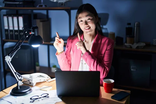 Chinese young woman working at the office at night smiling and looking at the camera pointing with two hands and fingers to the side.
