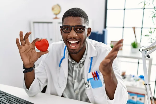 Young african american doctor man wearing doctor uniform holding heart at the clinic celebrating achievement with happy smile and winner expression with raised hand