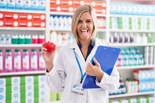 Young woman working at pharmacy drugstore holding heart sticking tongue out happy with funny expression.