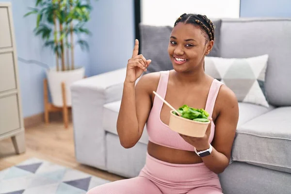 African american woman with braids eating salad after working out at home smiling with an idea or question pointing finger with happy face, number one