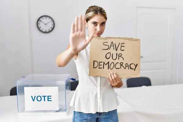 Young blonde woman at political election holding save out democracy banner with open hand doing stop sign with serious and confident expression, defense gesture