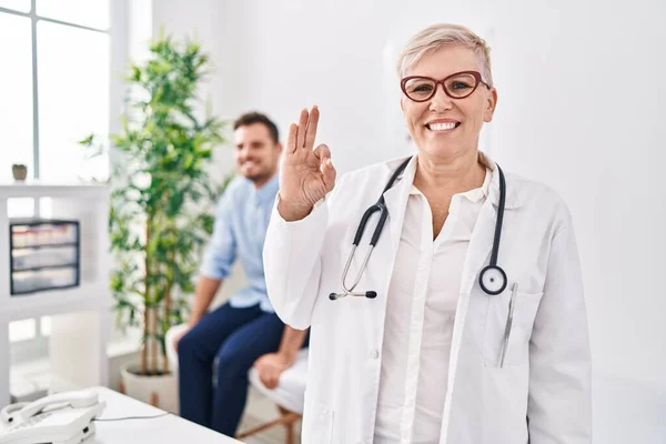 Female doctor wearing uniform and stethoscope at medical clinic doing ok sign with fingers, smiling friendly gesturing excellent symbol