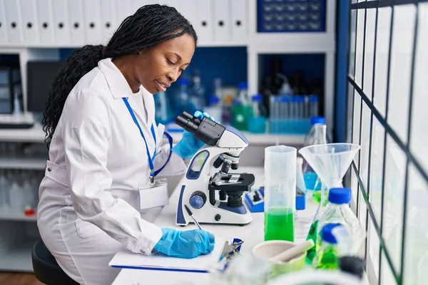 African American Woman Wearing Scientist Uniform Writing Clipboard Using Microscope — ストック写真