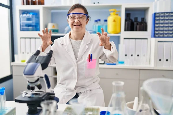 Hispanic girl with down syndrome working at scientist laboratory smiling funny doing claw gesture as cat, aggressive and sexy expression