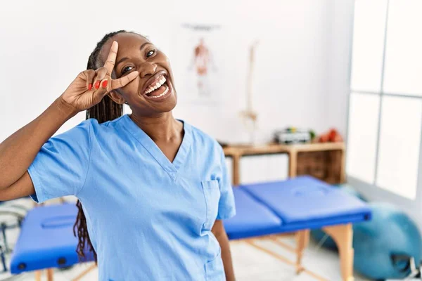 Black woman with braids working at pain recovery clinic doing peace symbol with fingers over face, smiling cheerful showing victory