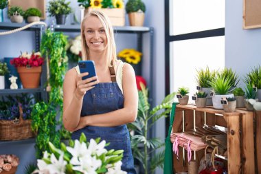 Young blonde woman florist smiling confident using smartphone at florist store