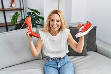 Beautiful blonde woman choosing high heel shoes and sneakers at home winking looking at the camera with sexy expression, cheerful and happy face. 