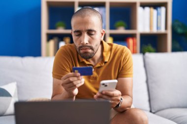 Young latin man using smartphone and credit card sitting on sofa at home