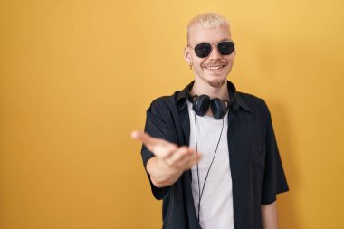 Sarı arka planda duran, güneş gözlüğü takan, neşeli gülümseyen, yardım eli uzatan ve kabul gören beyaz bir genç.. 