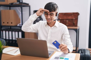 Young hispanic man working using computer laptop holding credit card very happy and smiling looking far away with hand over head. searching concept. 