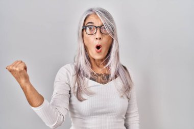 Beyaz arka planda duran gri saçlı orta yaşlı kadın elleri yana dönük, ağzı açık şaşkın bir ifadeyle şaşırdı.. 