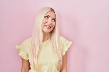 Caucasian woman standing over pink background looking away to side with smile on face, natural expression. laughing confident. 