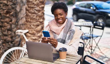 Laptop ve akıllı telefon kullanan Afro-Amerikalı bir kadın kahve dükkanının terasında oturuyor.