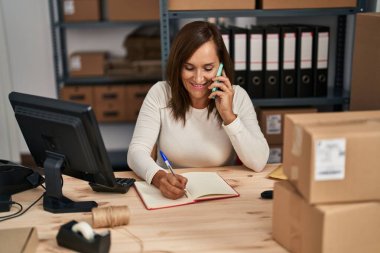 Middle age woman ecommerce business worker talking on the smartphone writing on book at office