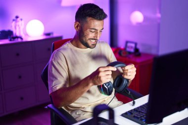 Oyun odasında kulaklık tutarak gülümseyen İspanyol asıllı genç adam.