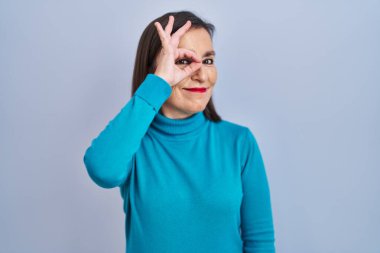 Orta yaşlı İspanyol bir kadın tecrit edilmiş arka planda duruyor. El hareketleriyle gülümsüyor, gözleriyle mutlu yüzle parmaklarının arasından bakıyor.. 