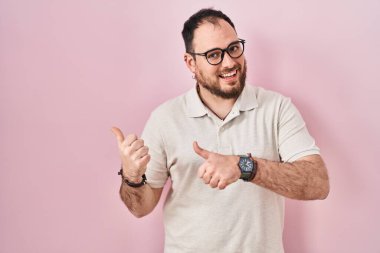 Plus size hispanic man with beard standing over pink background pointing to the back behind with hand and thumbs up, smiling confident 