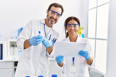 Orta yaşlı erkek ve kadın partnerler bilim adamı üniforması giyip doküman okuyor ve laboratuarda test tüpü tutuyorlar.