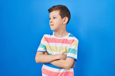 Mavi arka plan üzerinde duran genç beyaz çocuk kollarını kavuşturmuş, ikna olmuş ve kendinden emin bir şekilde yan tarafa bakıyor. 