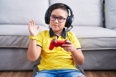 Young hispanic kid playing video game holding controller wearing headphones waiving saying hello happy and smiling, friendly welcome gesture 