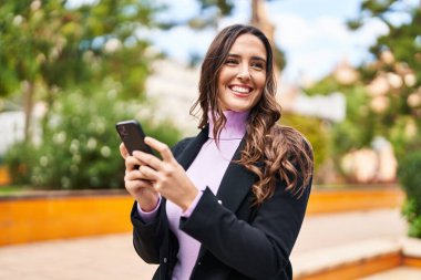 Genç İspanyol kadın parkta akıllı telefon kullanarak gülümsüyor.