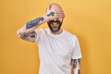 Sarı arka planda dikilen dövmeleri olan genç İspanyol bir adam. Gülümsüyor ve gülümsüyor. Gözleri şaşkınlıkla örtüyor. kör kavram. 