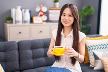 Genç Çinli kadın evdeki kanepede oturup kahve içiyor.